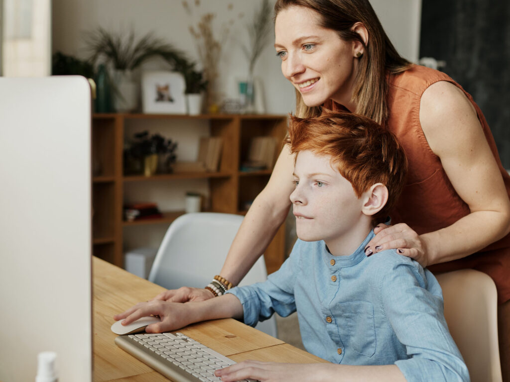 Mother with red hair in brown shirt teaching son how to a desktop on pine desk safely online 