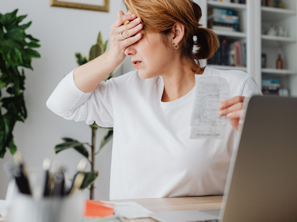 Mother in white shirt sitting at a desk looking shocked at laptop whilst holding financial documents.