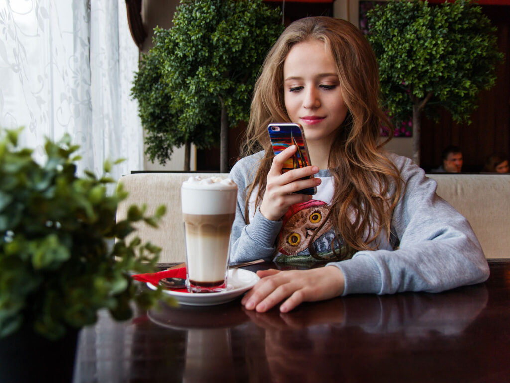 Teenager sitting in coffee shop with Latte whilst looking at their mobile phone