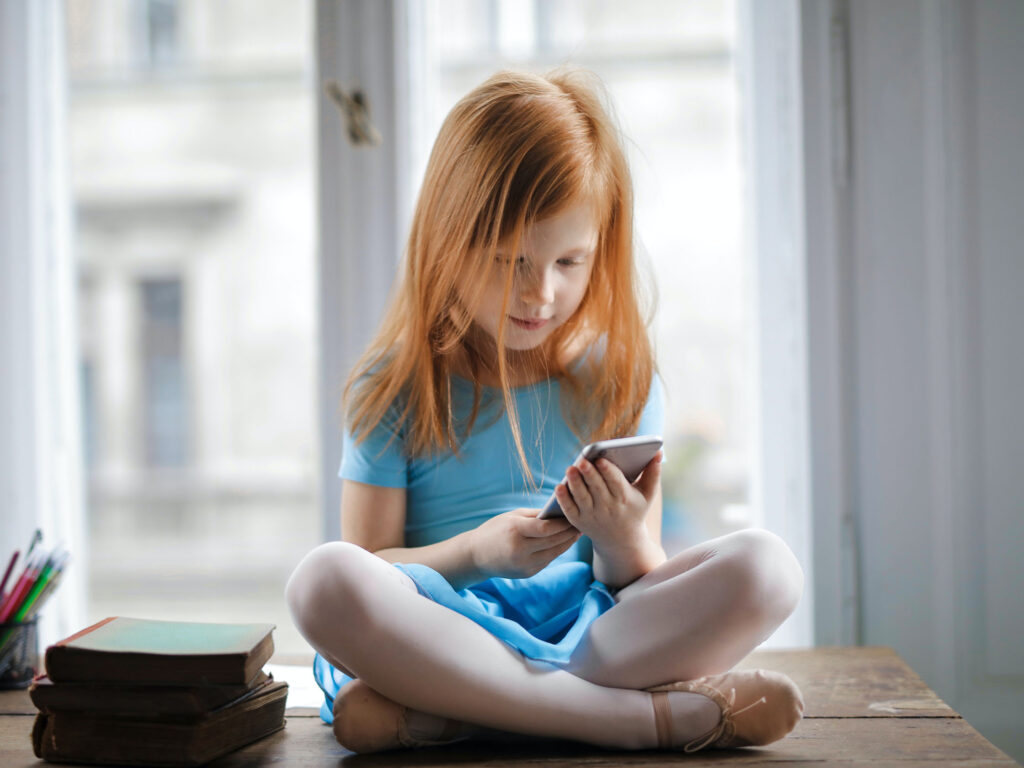 Young girl with red hair sitting on and oak table playing with what is assumed to be an android device