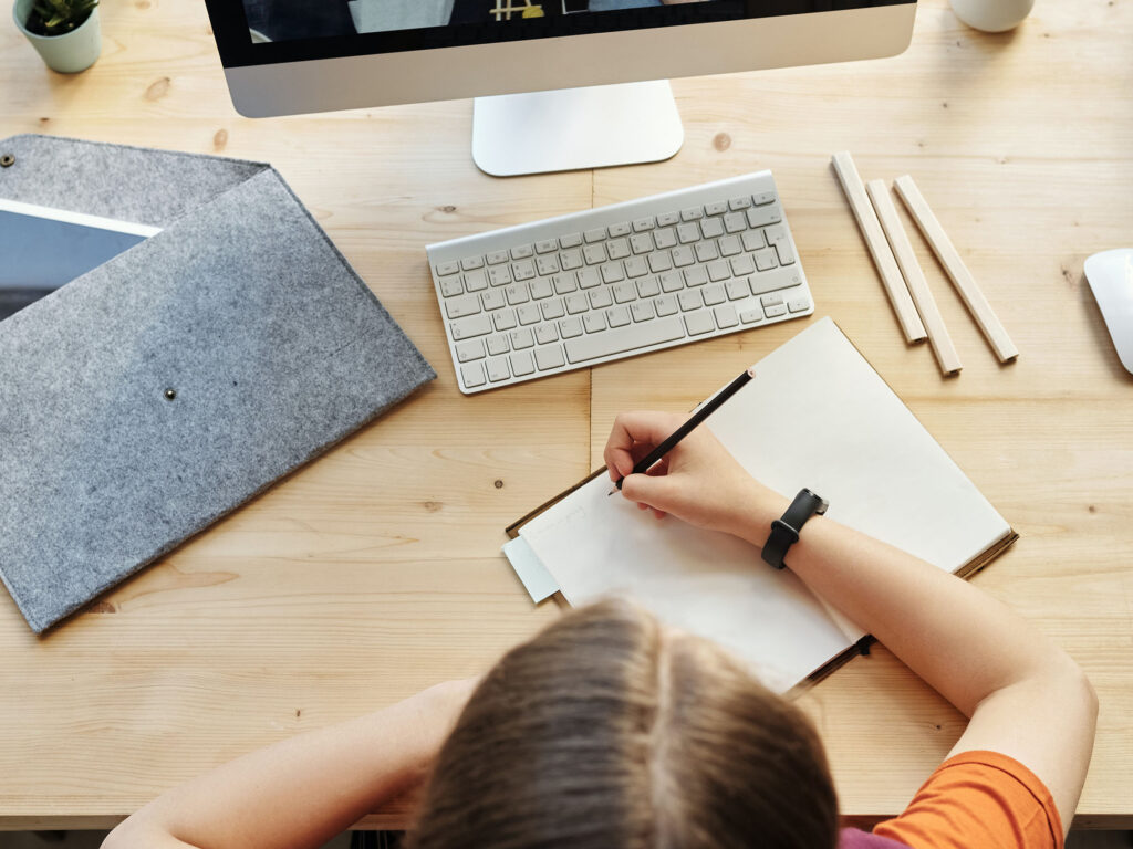 Teenager sitting down making notes at a pine desk in front of desktop computer screen